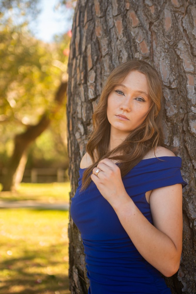 Chloe Dancey, portrait against a pine trunk in a royal blue off-shoulder dress